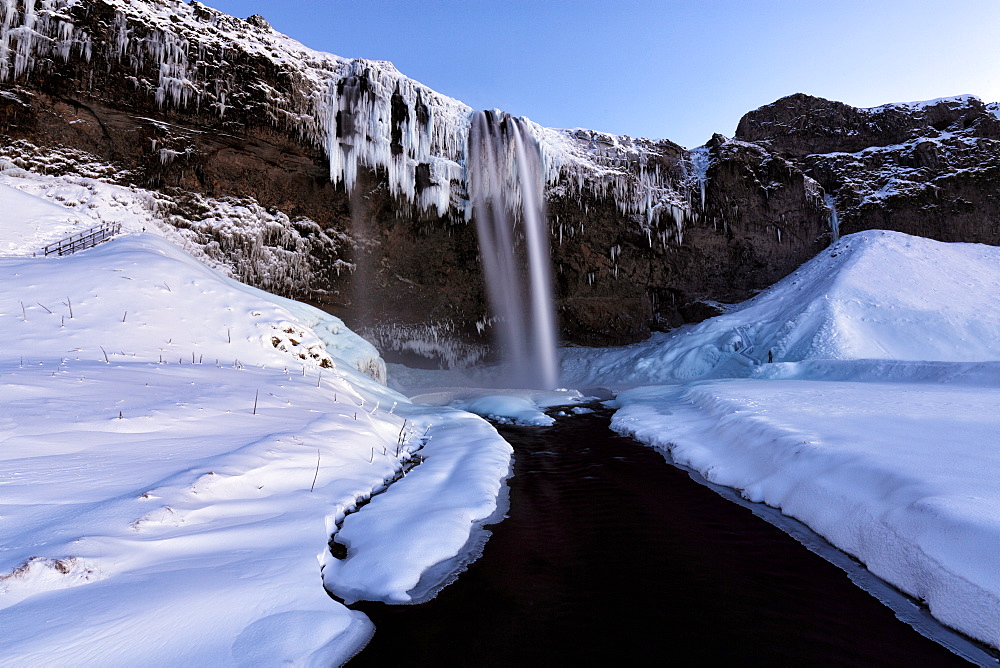 Winter view of Seljalandsfoss Waterfall at dusk with snow covered foreground and icicles hanging from cliffs, South Iceland, Polar Regions