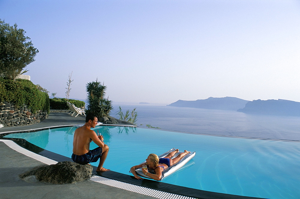 Holidaymakers on lilos in pool and view across bay, Oia, Santorini (Thira), Greek Islands, Greece, Europe