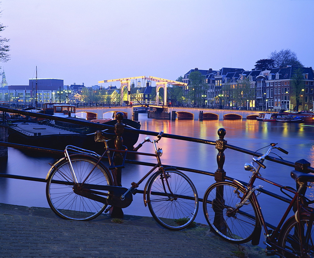 Bicycles by the side of the canal, with barges, and the Magere Brug (Skinny Bridge) in the background, in the evening in Amsterdam, Holland, Europe