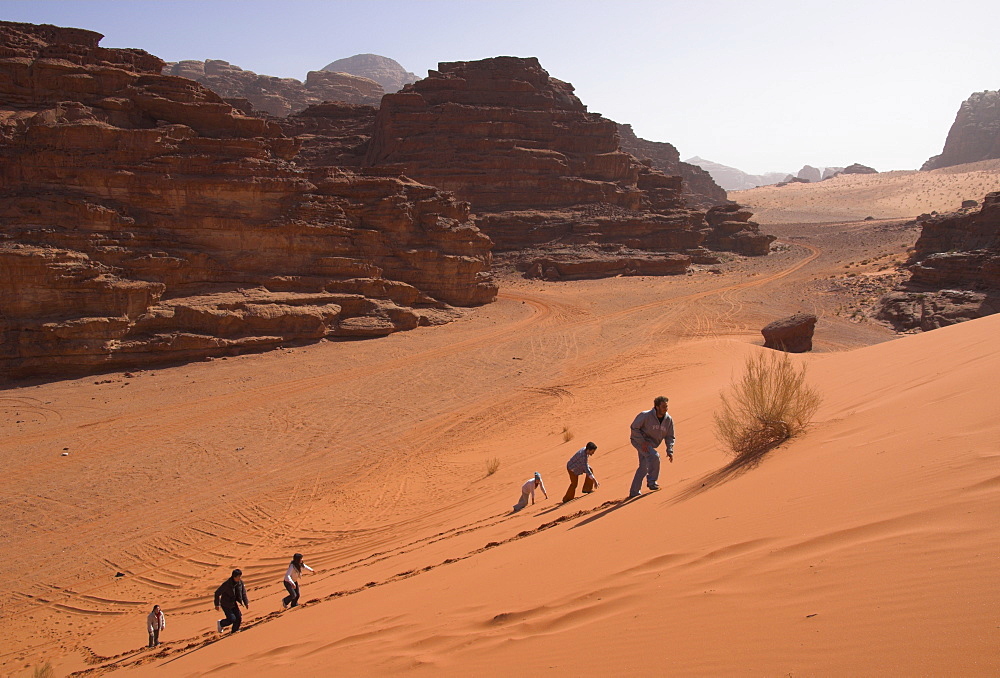 Young people climbing a typical red sand dune with rocks in background, Wadi Rum, Jordan, Middle East