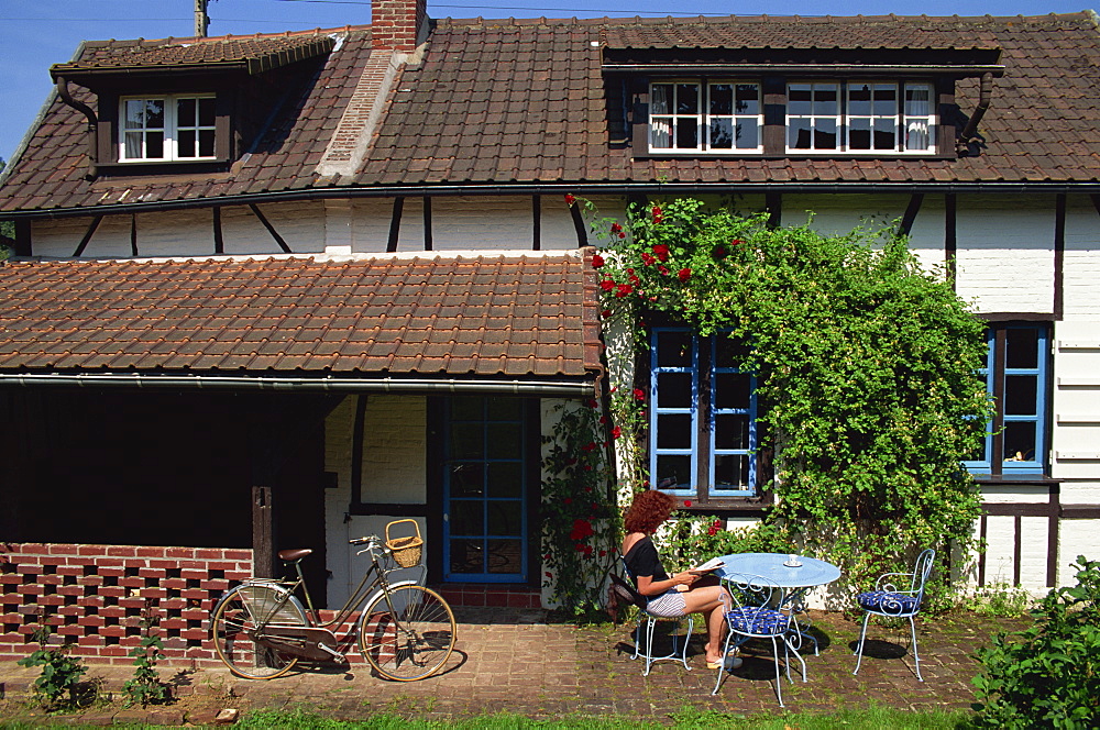 Woman reading at a table outside a typical house, with a bicycle in front, in Haute Normandie (Normandy), France, Europe