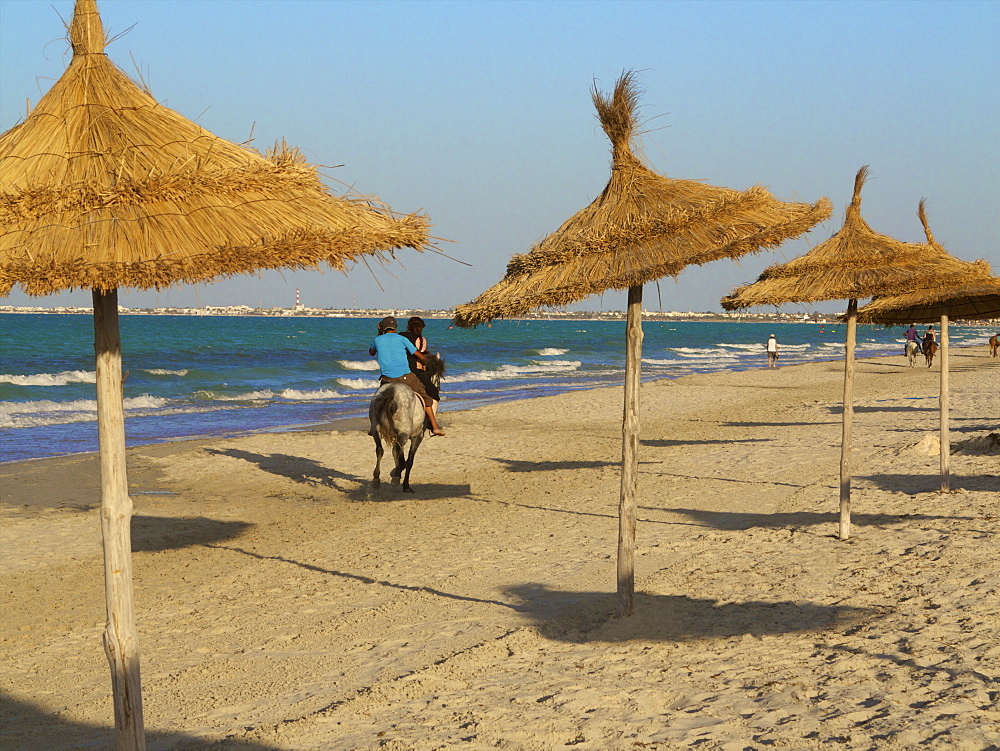 Tourists riding horses on the beach, with palm sunshades, at sunset, Djerba, Tunisia, North Africa, Africa