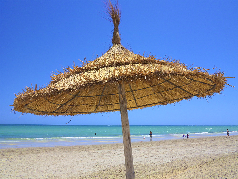 Palm sunshade on the sandy beach, and tourists walking along the sea, Djerba, Tunisia, North Africa, Africa
