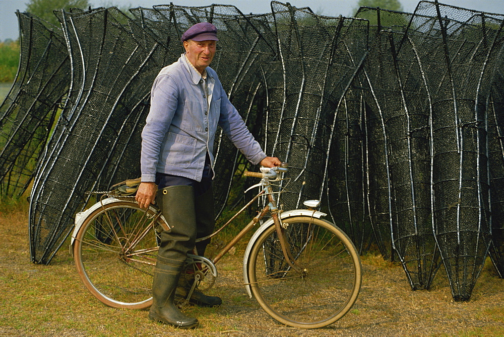 Portrait of a French eel fisherman on his bicycle looking at the camera, with fish traps behind, at the Lac de Grand Lieu in Western Loire, France, Europe