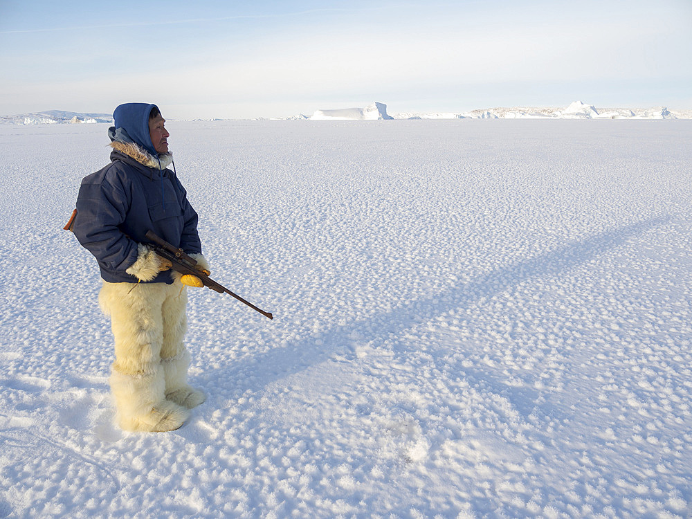 Watching a breathing hole of a seal. Inuit hunter wearing traditional trousers and boots made from polar bear fur on the sea ice of the Melville Bay near Kullorsuaq in North Greenland. North America, danish teritorry