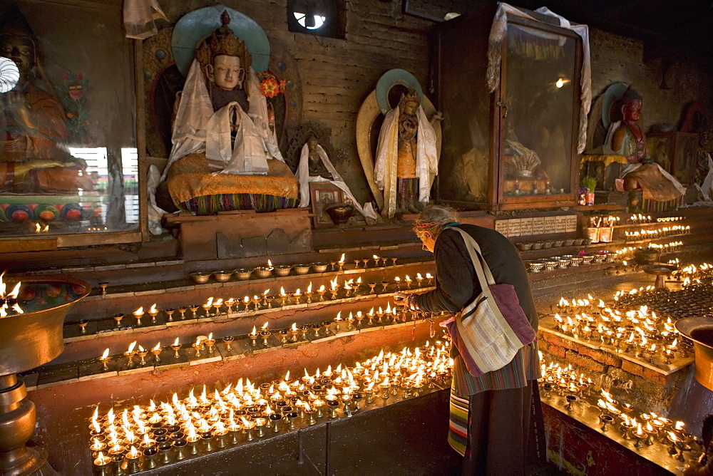 Old woman lighting butter lamps in front of Buddha statues draped with new silk scarves, in a small temple, at the Boudha or Bodhnath stupa, Tibetan New Year (Lhosar), Kathmandu, Nepal, Asia