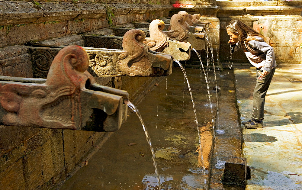 Girl takes a drink from the water spouts in a temple courtyard at Godavari in the south of the Kathmandu valley, Nepal, Asia