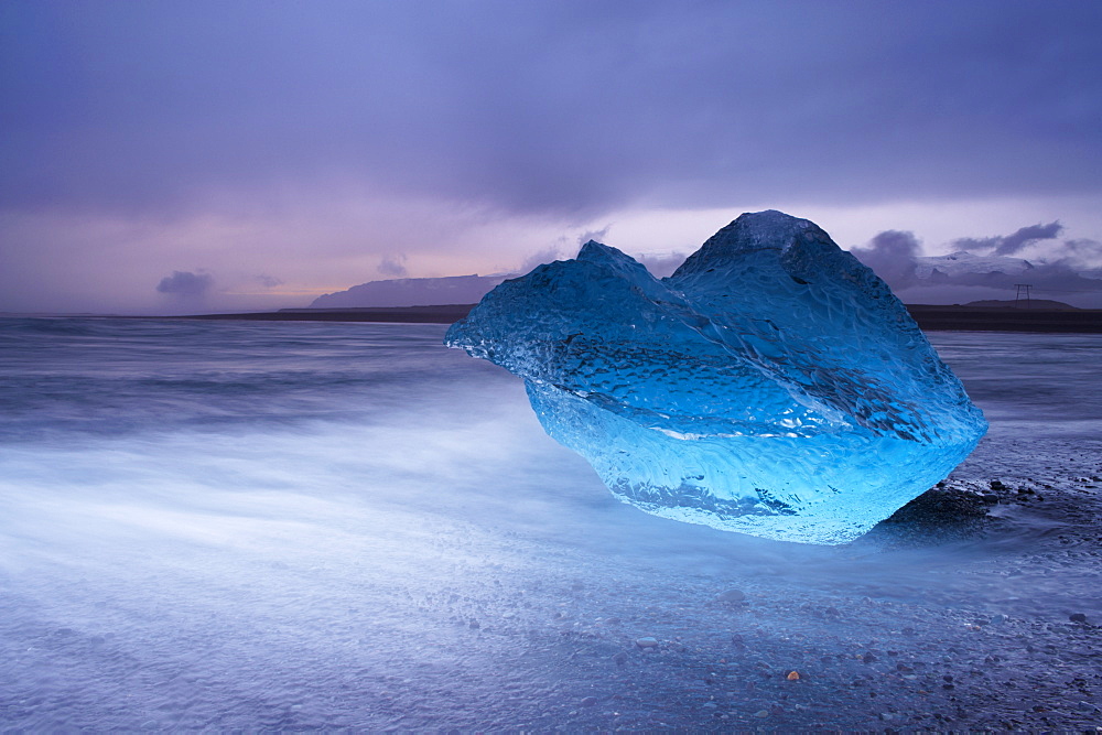 Translucent blue iceberg washed ashore on Breidamerkursandur black sands, near Jokulsarlon glacial lagoon, Oraefajokull (Vatnajokull) glacier in the distance, East Iceland, Polar Regions