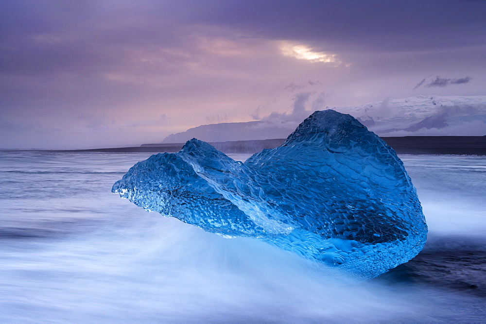 Translucent blue iceberg washed ashore on Breidamerkursandur black sands, near Jokulsarlon glacial lagoon, Oraefajokull (Vatnajokull) glacier in the distance, East Iceland, Polar Regions