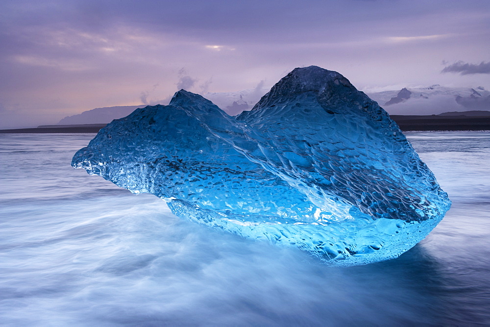 Translucent blue iceberg washed ashore on Breidamerkursandur black sands, near Jokulsarlon glacial lagoon, Oraefajokull (Vatnajokull) glacier in the distance, East Iceland, Polar Regions