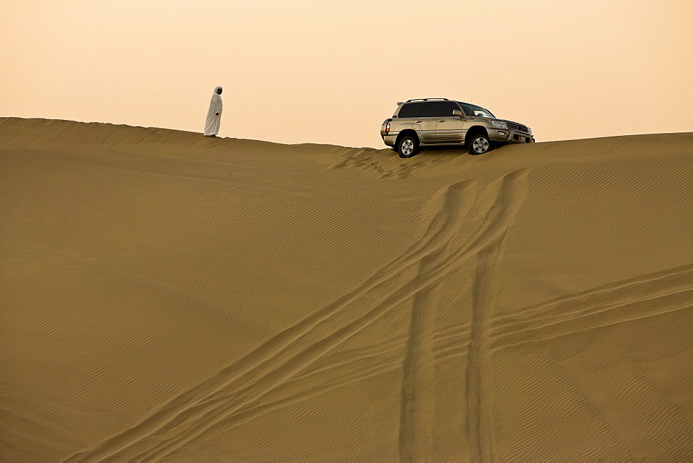 Man in traditional Arab dress looking at 4x4 car stuck on the ridge of a dune during a desert safari near Abu Dhabi, United Arab Emirates, Middle East