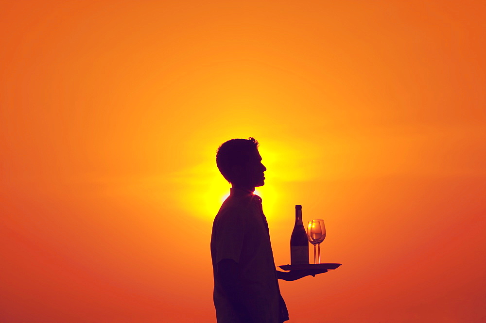 Waiter holding tray with bottle of wine and two glasses at sunset