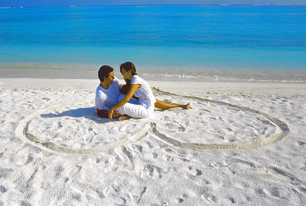 Young couple on beach sitting in a heart shaped imprint on the sand, Maldives, Indian Ocean, Asia