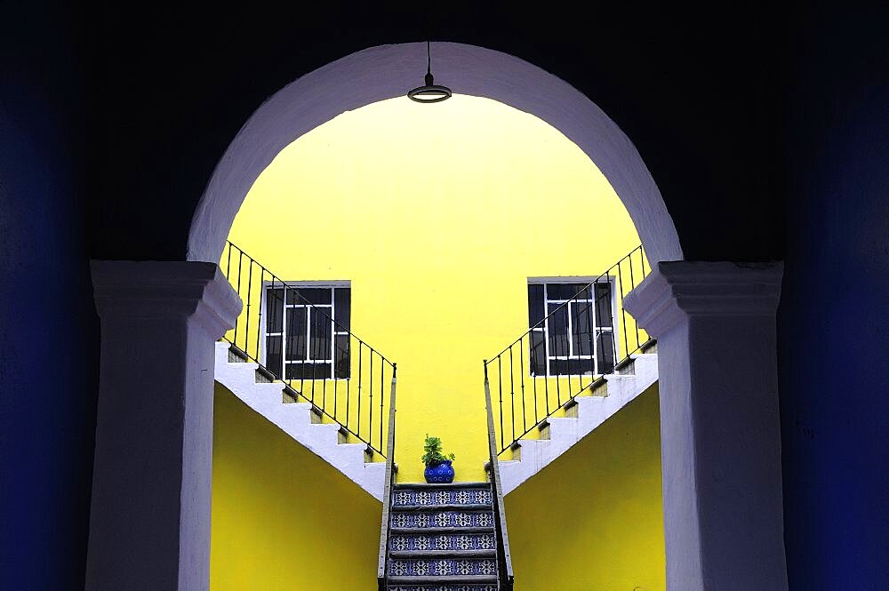 Mexico, Puebla, Looking into inner courtyard through archway in shadow towards yellow painted wall with blue and white tiled steps plant in blue pot and pair of square recessed windows.