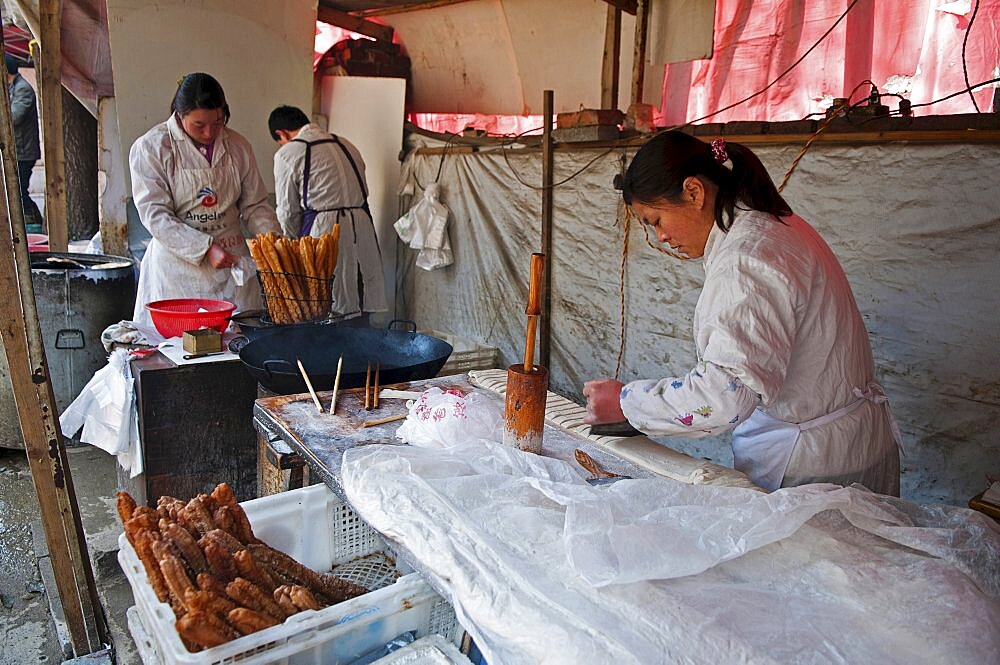 China, Jiangsu, Nanjing, Road-side bakery shop baker cutting strips of dough that will be twisted and deep-fried in a wok to ptoduce Yu Tiao a popular breakfast bread usually eaten with warm soyamilk or porridge or Xi Fan Bundles of Yu Tiao in baskets ready for sale.