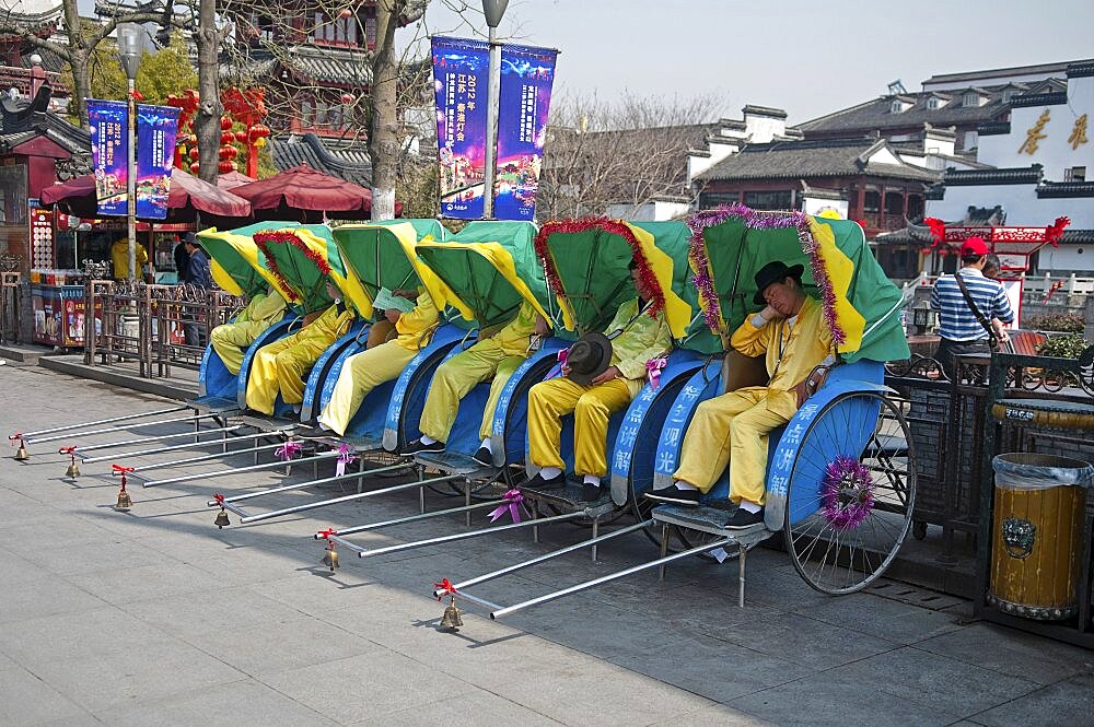 China, Jiangsu, Nanjing, Row of rickshaws with the rickshaw pullers in yellow uniform and hats asleep outside the Fuzi Temple a top tourist spot Fuzi Temple complex tile roofs and red Chinese lanterns in the background.