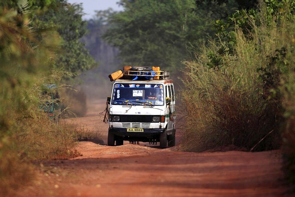 Gambia, Transport, Minibus driving down narrow dirt road covered in red soil carrying provisions on roof rack.