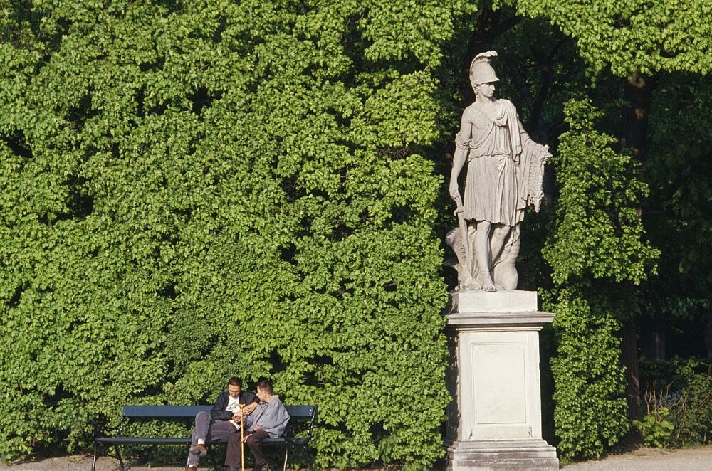 AUSTRIA  Vienna Schonbrunn Gardens.  Classical statue against hedge with two visitors sitting on bench at one side.