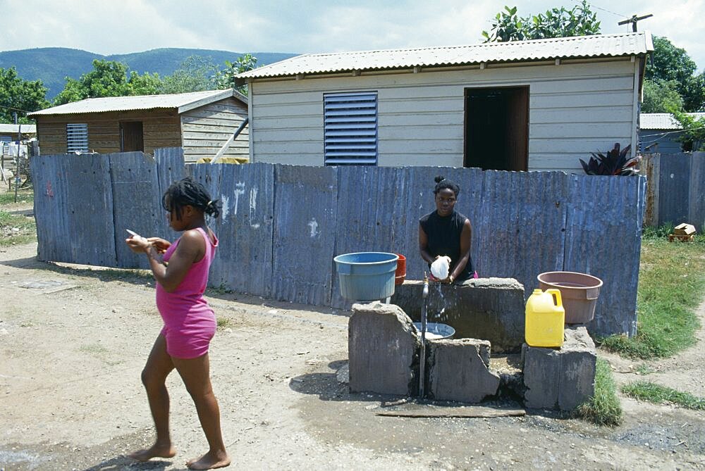 WEST INDIES Jamaica Kingston Bower Bank Transit Camp.  Girls beside water pipe with simple housing behind.