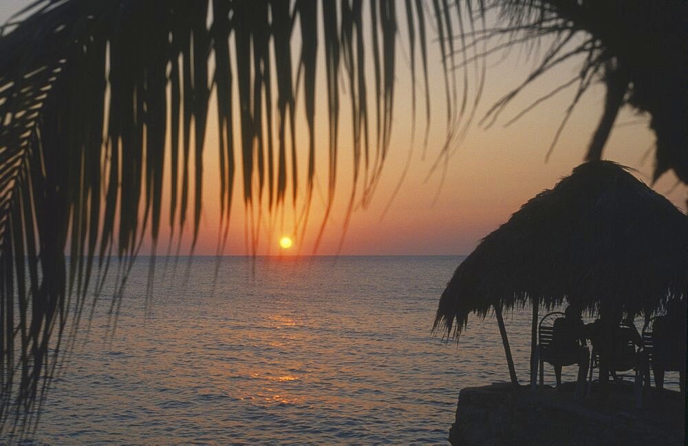 WEST INDIES Jamaica Negril Ricks Cafe at sunset through coconut palm tree with tourist sitting under thatched sun shade