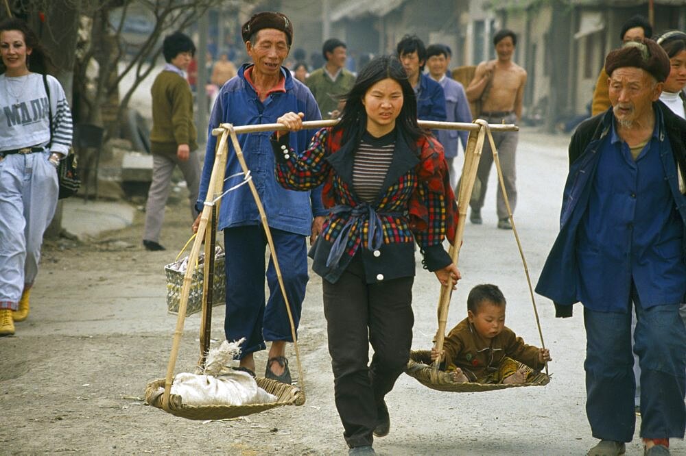 CHINA Bijie Woman with carrying scales over her shoulders balancing a small child with a sack.