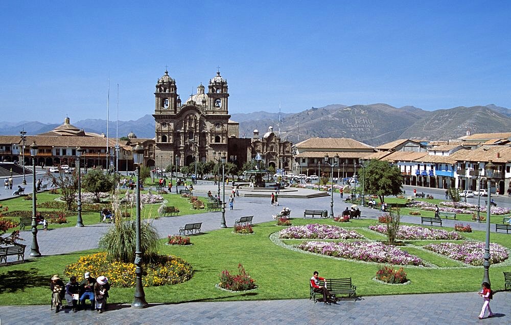 PERU  Cusco Looking across Plaza de Armas to Iglesia La Compania de Jesus. Cuzco Travel Tourism Holiday Vacation Explore Recreation Leisure Sightseeing Tourist Attraction Tour Destination Plaza De Armas Cusco Cuzco Peru Peruvian South Southern America Latin Sacred Valley Of The Inca Incas Inka Inkas Incan Vivid Vibrant City Town Square Tranquil Tranquility Tradition Traditional Culture Cultural Buildings Architecture Architectural Ethnic UNESCO World Heritage Site Panorama Panoramic Andes Andean Hills Hillside Mountain Range Mountainous View Vista Spanish Colonial Scenic Picturesque Backdrop People Visitors Pavement Sidewalk Park Garden Path Pathway Centre Iglesia La Compania Jesus Church Religion Religious Jesuit Worship Spectacular Beautiful Peaceful Quiet Blue Sky Magnificent Christianity Catholic Surrounded Calendar American Center South America