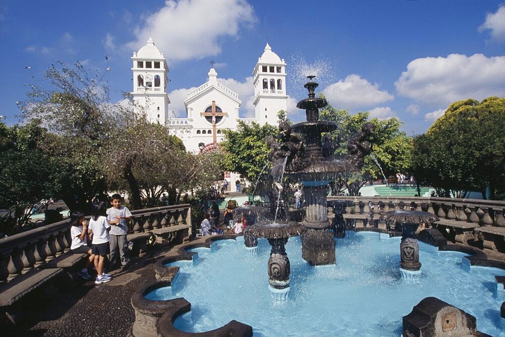 EL SALVADOR  Juayua Temple del Senor de Juayua and Plaza.  Children beside fountain in foreground with white exterior facade of the Church of the Cristo Negro or Black Christ behind. American Kids Religion Religious Immature