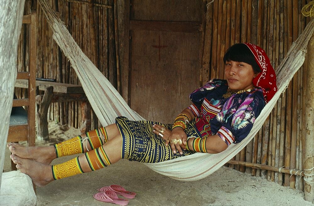 PANAMA San Blas Islands Kuna Indians Young Kuna mother rests in hammock outside home  wearing brightly coloured traditional layered applique mola panel    traditional bead design arm and leg bands and gold nose ring. Cuna Caribbean American Central America Classic Classical Colored Female Women Girl Lady Hispanic Historical Latin America Latino Older Panamanian West Indies