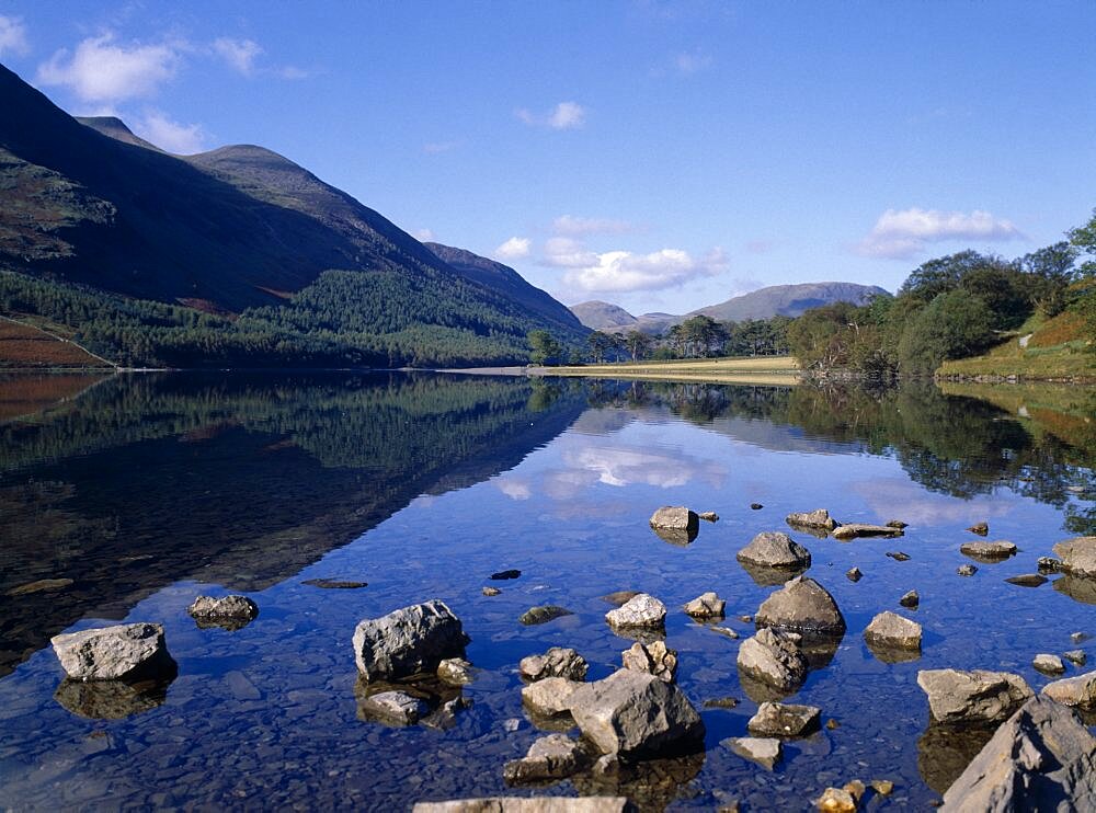 Buttermere, Landscape blue sky and drifting cloud reflected in lake with small rocks protruding from clear shallow water in the foreground, Lake District, Cumbria, England, United Kingdom