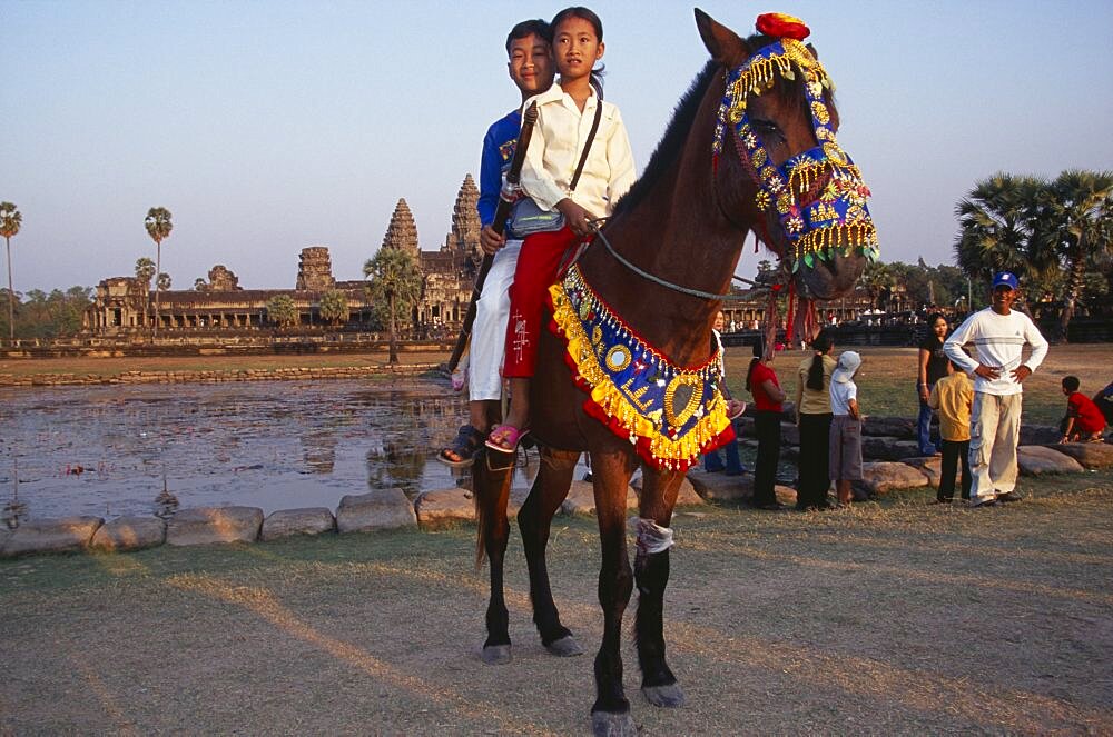 Cambodian children visiting Angkor during the Chinese New Year having photographs taken on pony wearing decorative harness in front of lake and temple complex, Angkor Wat, Siem Reap Province, Cambodia