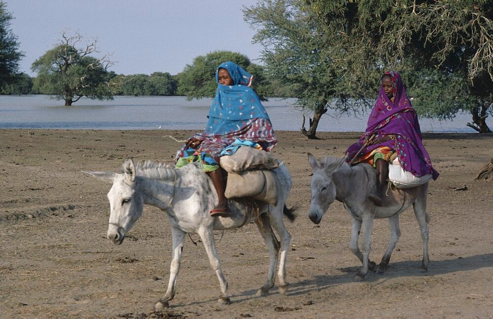 Baggara Arabs, Two women from the Beni Halba tribe riding donkeys past lake, South Darfur, Sudan