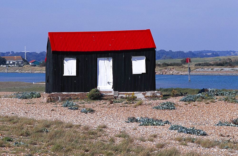 Rye harbour, Red and black corrugated hut with white doors built on the shingle with the harbour inlet behind, Rye, East sussex, England, United Kingdom