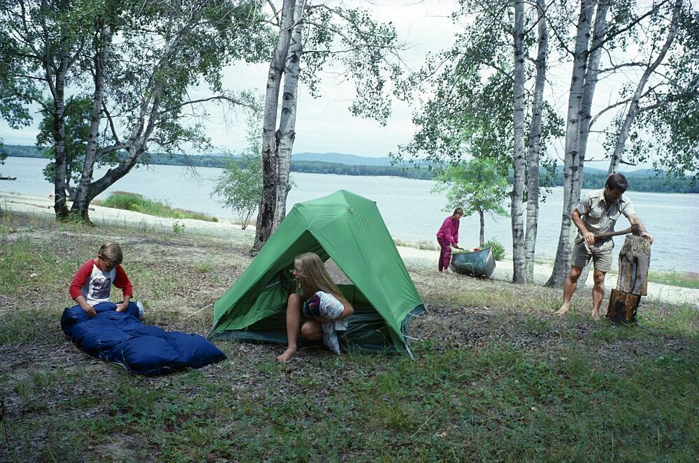 Canadian family on outback holiday setting up camp beside lake, Children with tent and sleeping bag mother with canoe and father using axe to split log, Camping, Canada