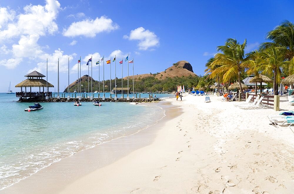 Tourists on the beach at Sandals Grande St Lucian Spa and Beach Resort hotel beside a wooden jetty with Pigeon Island National Historic Park beyond, Gros Islet, St Lucia