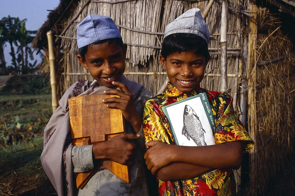 Three-quarter portrait of two smiling young boys carrying books and writing board standing outside thatched hut, Bangladesh, Asia