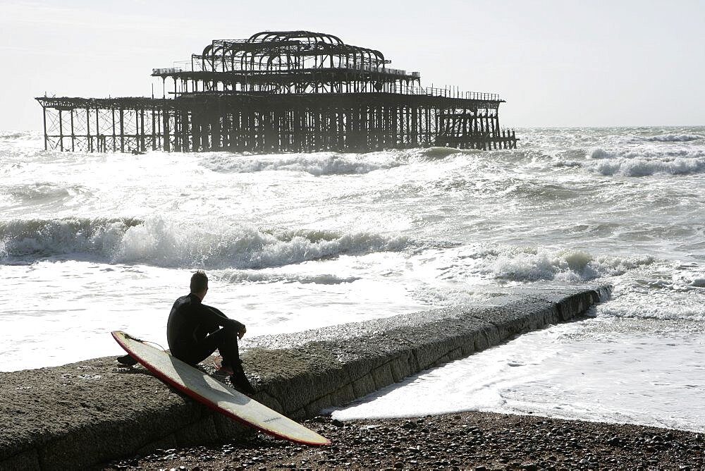 England, East Sussex, Brighton, man sat with surf board on goyne in front of the burnt out shell of the former West Pier.