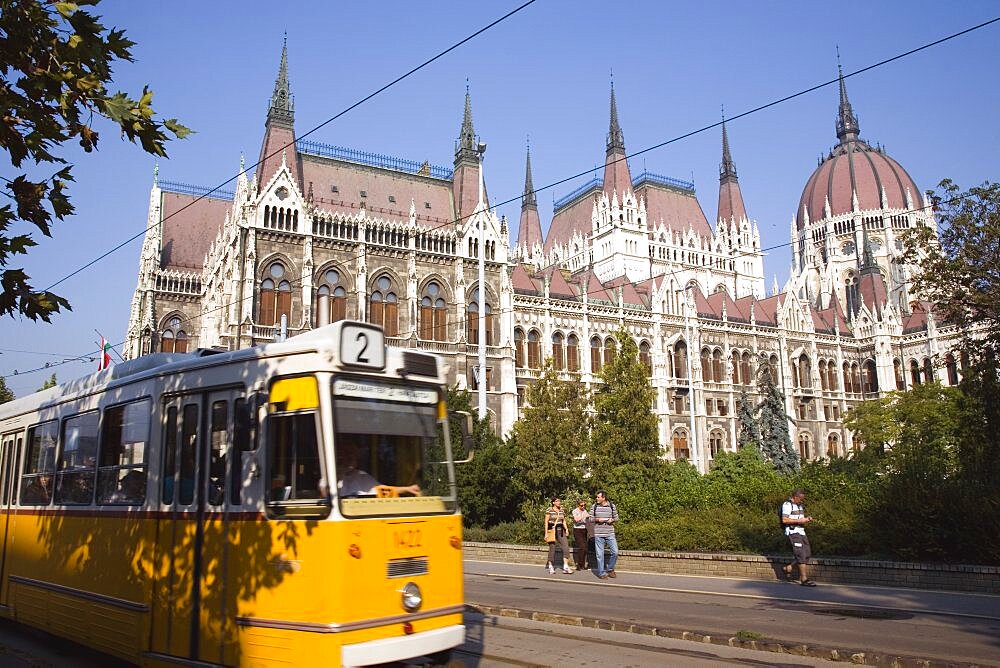 Hungary, Pest County, Budapest, yellow tram passing the Neo-Gothic exterior facade of the Parliament Building with pedestrians on pavement beyond tram line