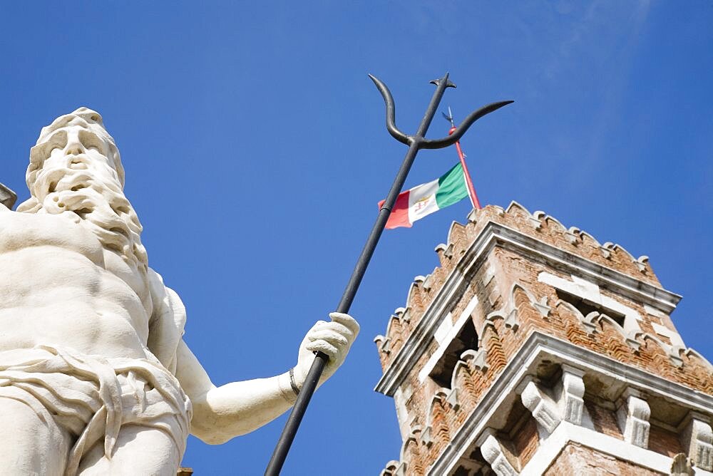 Italy, Veneto, Venice, Centro Storico, Arsenale, Part view of crenellated tower flying Italian tricolour flag with statue of Neptune in foreground against clear blue sky of late summer