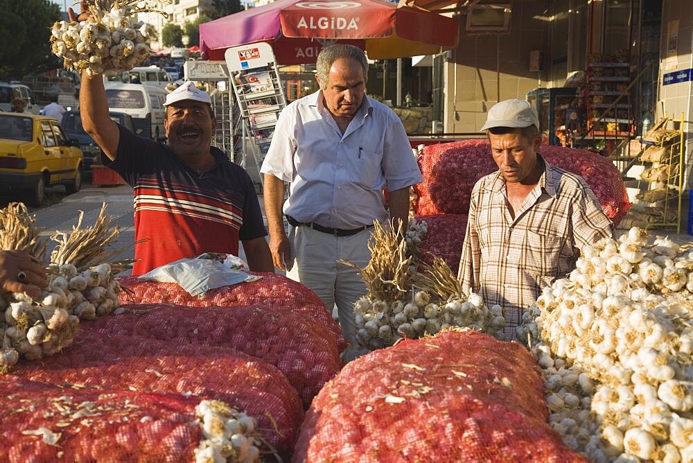 Turkey, Aydin Province, Kusadasi, Stallholder selling garlic at weekly market standing behind stall stacked high with sacks of garlic bulbs and holding up bunch of tied bulbs in late afternoon sunshine