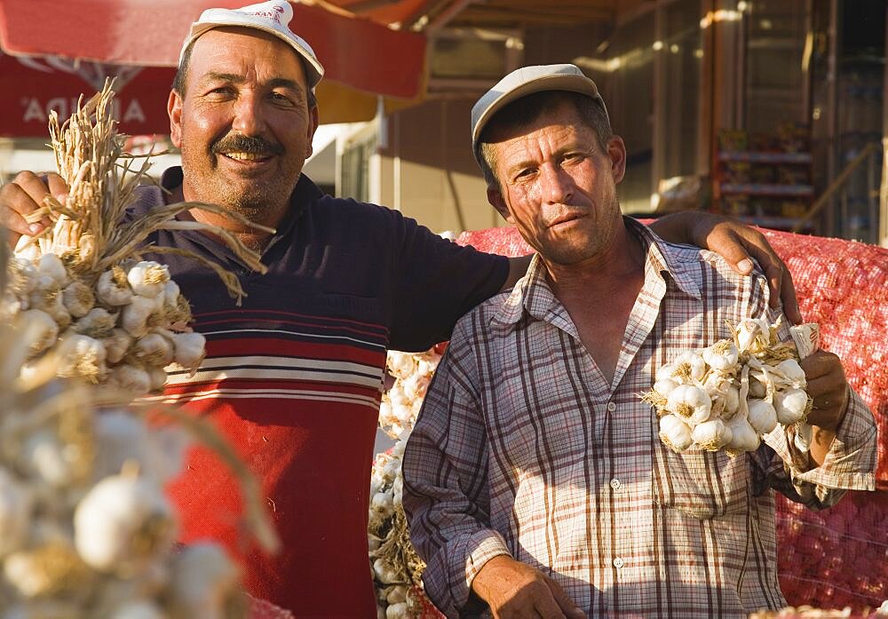 Turkey, Aydin Province, Kusadasi, Stallholders at weekly market with sacks of garlic for sale and holding up bunches of bulbs in late afternoon summer sunshine