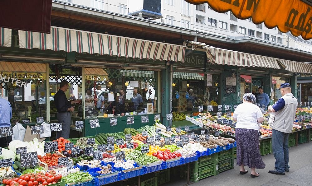 Austria, Vienna, The Naschmarkt, Customers at fresh produce stall with display including peppers, leeks, tomatoes and mushrooms in foreground of glass shopfronts