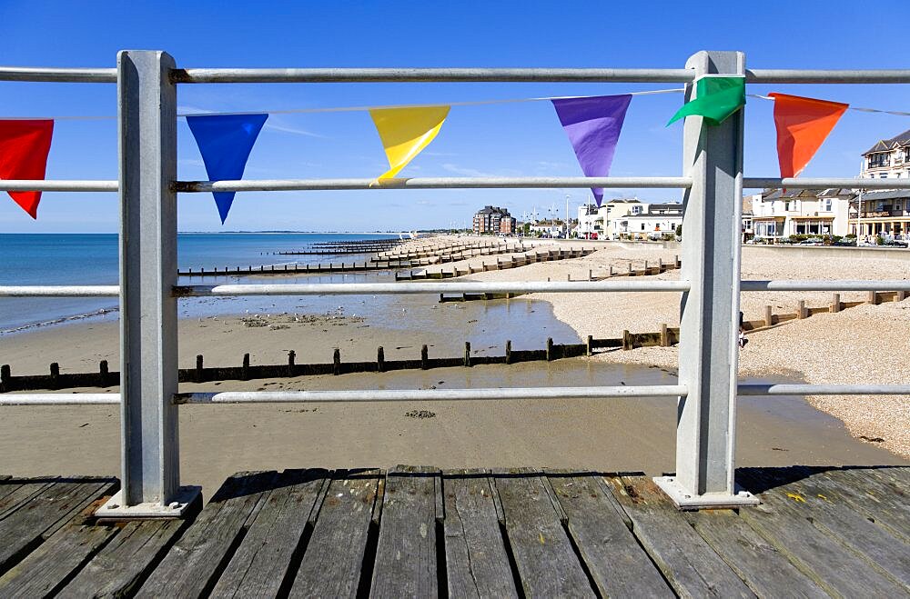 England, West Sussex; Bognor Regis, Wooden groynes at low tide used as sea defences against erosion of the shingle pebble beach seen through metal railings with colourful flags on the pier.