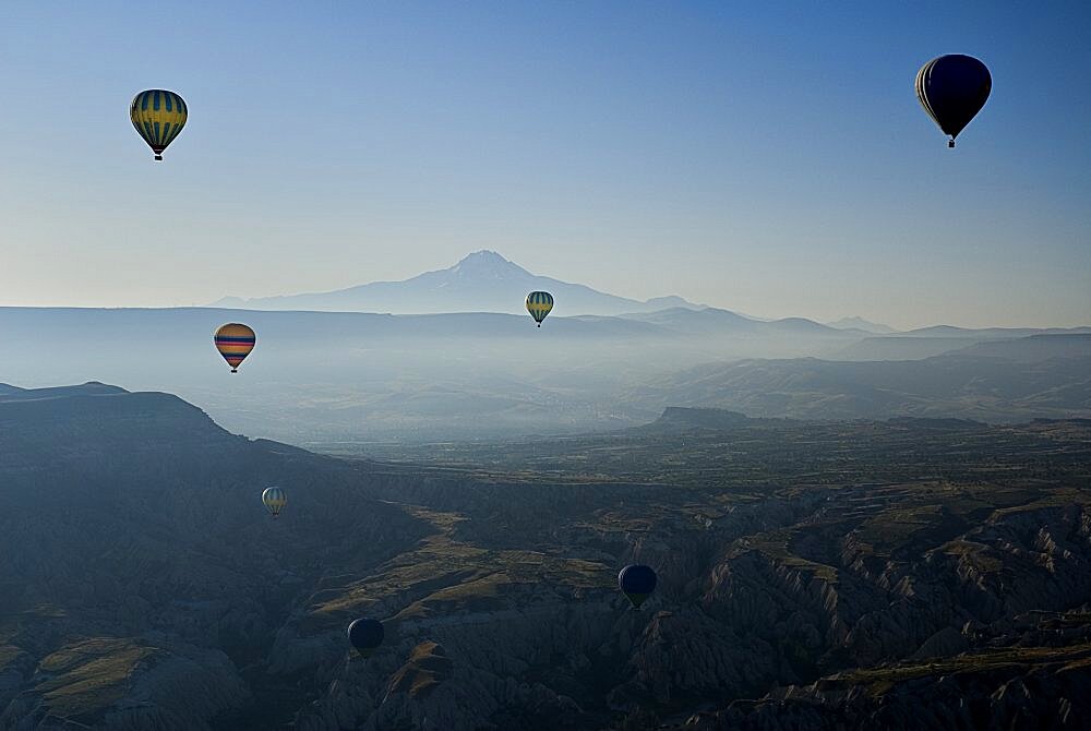 Turkey, Cappadocia, Goreme, Early morning with hot air balloons in flight mist drifting across landscape and Mount Erciyes in the background.