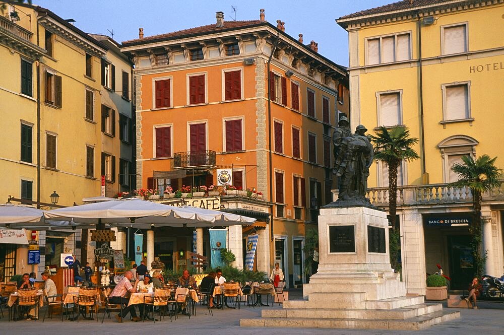 People sitting at outside cafe tables in piazza of town beside Lake Garda Facades of buildings bank and hotel painted pale yellow and orange with window shutters, Salo, Lombardy, Italy