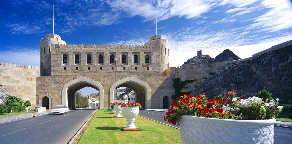 OMAN  Muscat Muscat Gate Museum   Crenellated gatehouse with archways over roads divided by central strip of grass with  white tubs of red and white flowers.