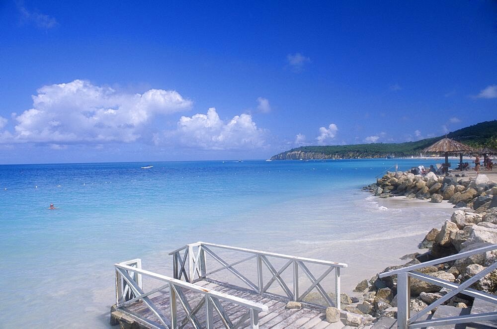 WEST INDIES Antigua Dickenson Bay Raised area of sand fortified by rocks with people sitting on loungers under thatched umbrella with man swimming and wooden jetty in the foreground.