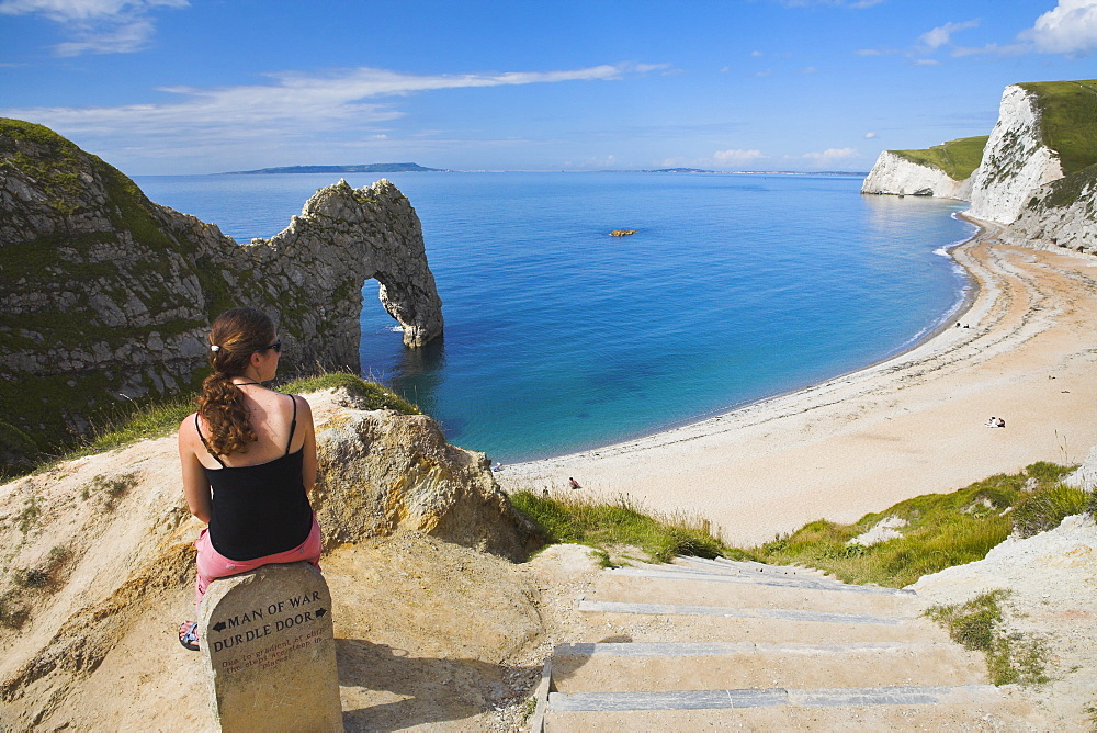 Woman resting on a clifftop sign, looking towards Durdle Door beach, Jurassic Coast, UNESCO World Heritage Site, Dorset, England, United Kingdom, Europe