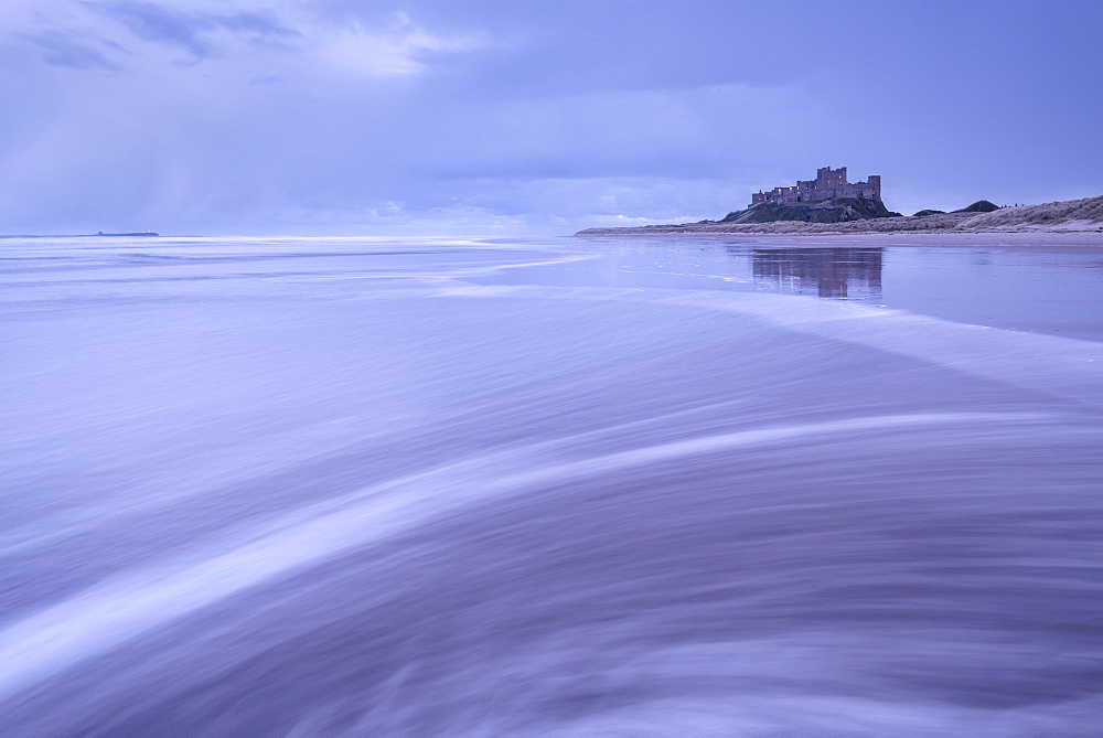 Waves wash over the beach next to Bamburgh Castle on a stormy winter evening, Northumberland, England, United Kingdom, Europe