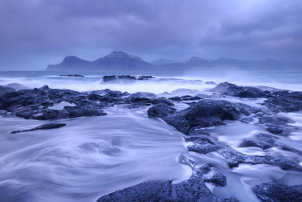 Waves wash over the dark basalt ledges of Gjogv on a stormy evening in the Faroe Islands, Denmark, Europe