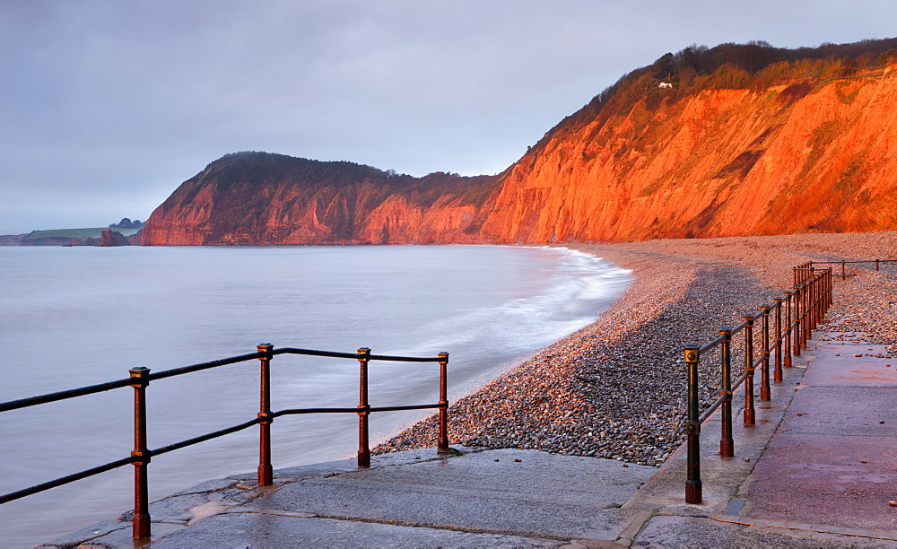 Early morning sunlight glows against the distinctive red cliffs of High Peak, viewed from the beach at Sidmouth, Devon, England, United Kingdom, Europe